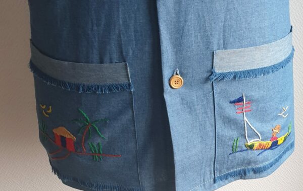 Chemise carabela traditionnelle haïtienne avec son col classique. Cinq boutons en bois sont disposés sur le devant en ligne centrale. Trois poches brodées à la main avec sur la poche droite une petite maison traditionnelle ancestrale. Une poche miniaturisée se distingue sur la demie manche gauche.