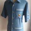 Chemise carabela traditionnelle haïtienne avec son col classique. Cinq boutons en bois sont disposés sur le devant en ligne centrale. Trois poches brodées à la main avec sur la poche droite une petite maison traditionnelle ancestrale. Une poche miniaturisée se distingue sur la demie manche gauche.