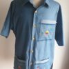 Chemise carabela traditionnelle haïtienne avec son col classique. Cinq boutons en bois sont disposés sur le devant en ligne centrale. Trois poches brodées à la main avec sur la poche droite une petite maison traditionnelle ancestrale. Une poche miniaturisée se distingue sur la demie manche gauche.