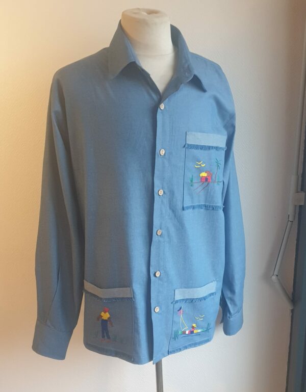 126.1Chemise carabela traditionnelle haïtienne de couleur bleue confectionnée en France. Trois poches brodées main. Ensemble de boutons verticaux et centraux en bois coco. Tissu cent pour cent coton. Création artisanale Anjero.