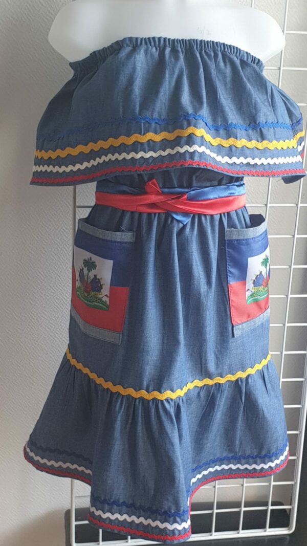 2.1Robe enfant carabela sur une tenue traditionnelle haïtienne. Un ruban élastique à la taille est intégré avec le placement de deux jolies poches l'une comme l'autre décorées avec le drapeau haïtien. Une ceinture bicolore est mise en évidence et singularise ce vêtement dont l'aisance et la douceur qui seront une révélation au porté, ne laisseront pas de vous surprendre. Taille : 6 et 7 ans. Prix : 109.95€. Cette confection est réalisée à Paris et est signée par le maître tailleur AnjeroCréation. 