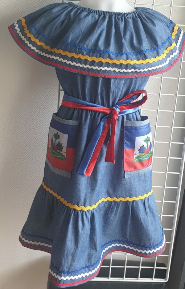2.2Robe enfant carabela sur une tenue traditionnelle haïtienne. Un ruban élastique à la taille est intégré avec le placement de deux jolies poches l'une comme l'autre décorées avec le drapeau haïtien. Une ceinture bicolore est mise en évidence et singularise ce vêtement dont l'aisance et la douceur qui seront une révélation au porté, ne laisseront pas de vous surprendre. Cette confection est réalisée à Paris et est signée par le maître tailleur AnjeroCréation.