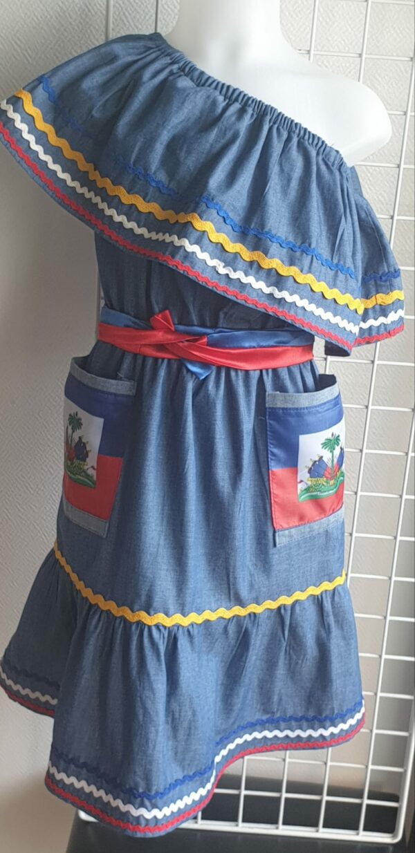 2.3Robe enfant carabela sur une tenue traditionnelle haïtienne. Un ruban élastique à la taille est intégré avec le placement de deux jolies poches l'une comme l'autre décorées avec le drapeau haïtien. Une ceinture bicolore est mise en évidence et singularise ce vêtement dont l'aisance et la douceur qui seront une révélation au porté, ne laisseront pas de vous surprendre. Cette confection est réalisée à Paris et est signée par le maître tailleur AnjeroCréation.