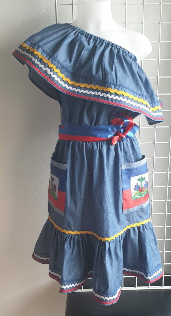 3.1Robe enfant carabela sur une tenue traditionnelle haïtienne. Un ruban élastique à la taille est intégré avec le placement de deux jolies poches l'une comme l'autre décorées avec le drapeau haïtien. Une ceinture bicolore est mise en évidence et singularise ce vêtement dont l'aisance et la douceur qui seront une révélation au porté, ne laisseront pas de vous surprendre. Taille : 9 et 10 ans. Prix : 125.95€. Cette confection est réalisée à Paris et est signée par le maître tailleur AnjeroCréation. 