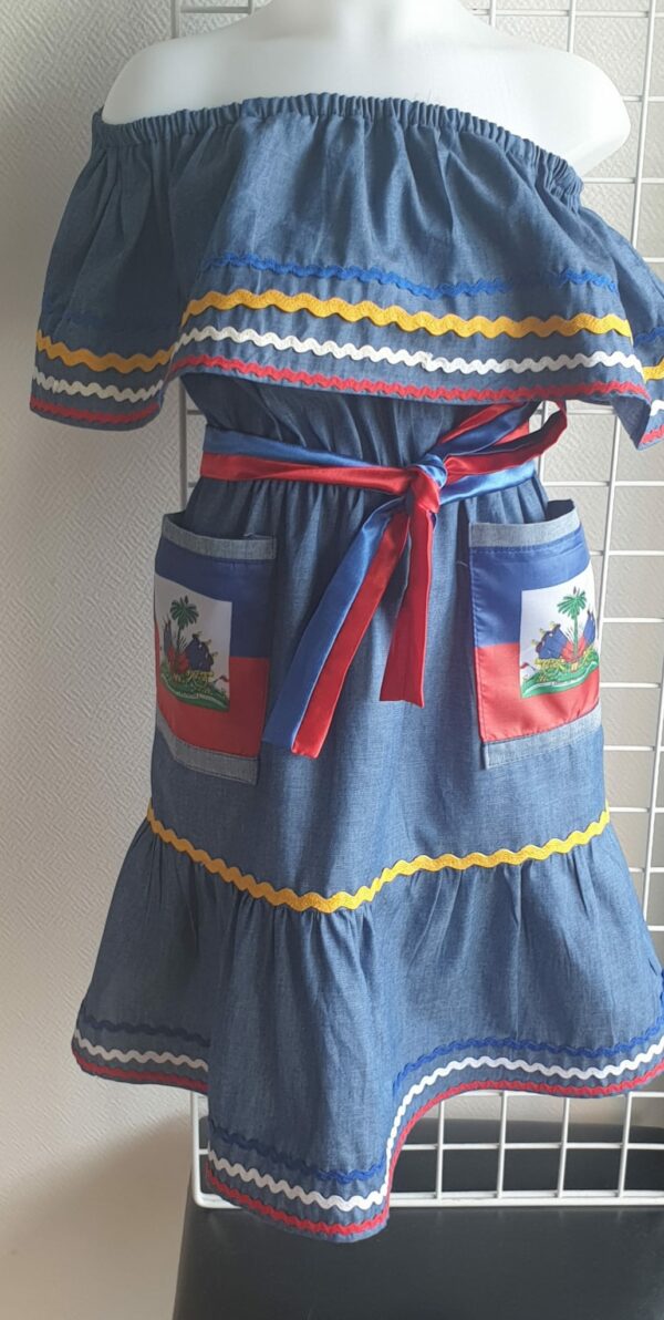 3.2Robe enfant carabela sur une tenue traditionnelle haïtienne. Un ruban élastique à la taille est intégré avec le placement de deux jolies poches l'une comme l'autre décorées avec le drapeau haïtien. Une ceinture bicolore est mise en évidence et singularise ce vêtement dont l'aisance et la douceur qui seront une révélation au porté, ne laisseront pas de vous surprendre. Cette confection est réalisée à Paris et est signée par le maître tailleur AnjeroCréation. 