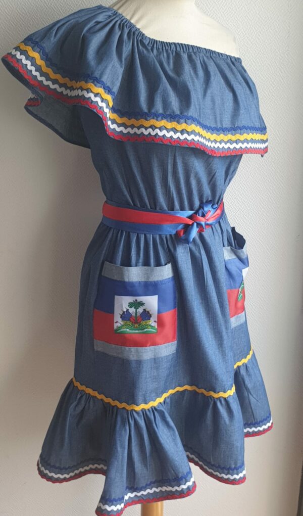 4.1Robe enfant carabela sur une tenue traditionnelle haïtienne. Ce vêtement est élastique à la taille. Il est présenté avec ses deux poches ornées du draapeau d'Haïti et sa ceinture bicolore en rouge et bleue. Taille : 6, 7 ans. Entièrement conçu et réalisé aux ateliers AnjeroCréation à Paris. Prix : 109.95€.