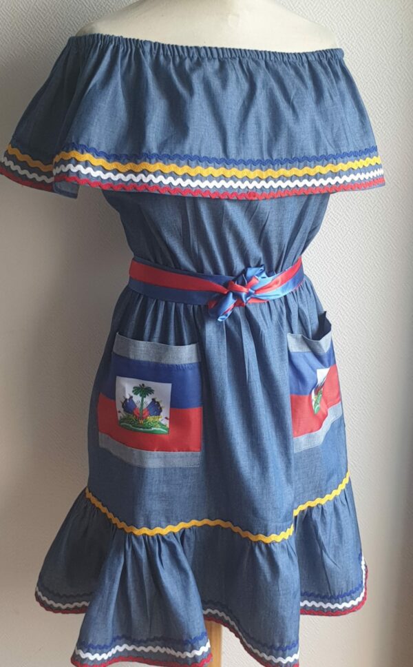 5.3Robe enfant carabela sur une tenue traditionnelle haïtienne en bleu clair. Ce vêtement est élastique à la taille. Il est présenté avec ses deux poches ornées du draapeau d'Haïti et sa ceinture bicolore en rouge et bleue. Entièrement conçu et réalisé aux ateliers AnjeroCréation à Paris.