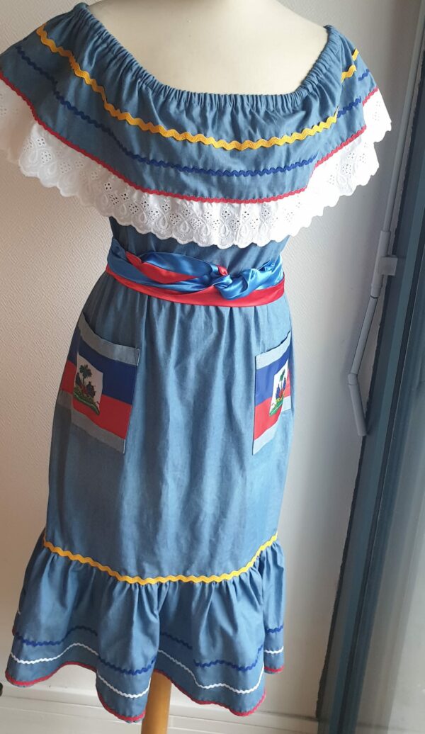 12.1Robe carabela en bleu clair sur une tenue traditionnelle haïtienne. Son élégance est mise en valeur par le tissage d'un col bardot en dentelles anglaises ainsi que par deux poches latérales symétriques . Des zigzags multicolors sur le haut et le bas de robe ajoutent à l'harmonie du vêtement doux et agréable à porter. Taille : XL. Entièrement confectionnée aux ateliers AnjéroCréation à Paris. Prix : 179.95€. 