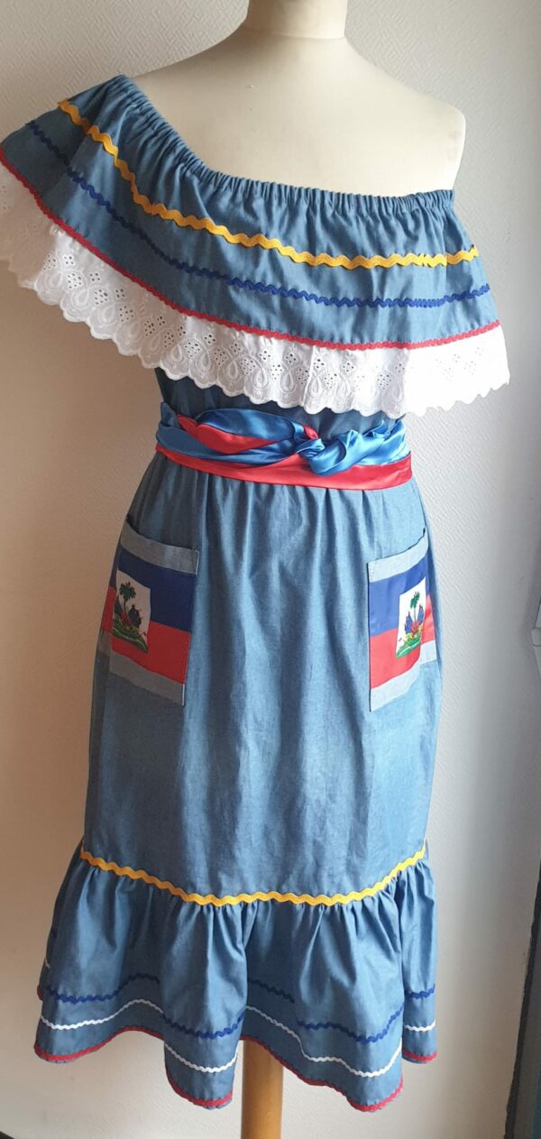 12.2Robe carabela en bleu clair sur une tenue traditionnelle haïtienne. Son élégance est mise en valeur par le tissage d'un col bardot en dentelles anglaises ainsi que par deux poches latérales symétriques . Des zigzags multicolors sur le haut et le bas de robe ajoutent à l'harmonie du vêtement doux et agréable à porter. Taille : XL. Entièrement confectionnée aux ateliers AnjéroCréation à Paris. 