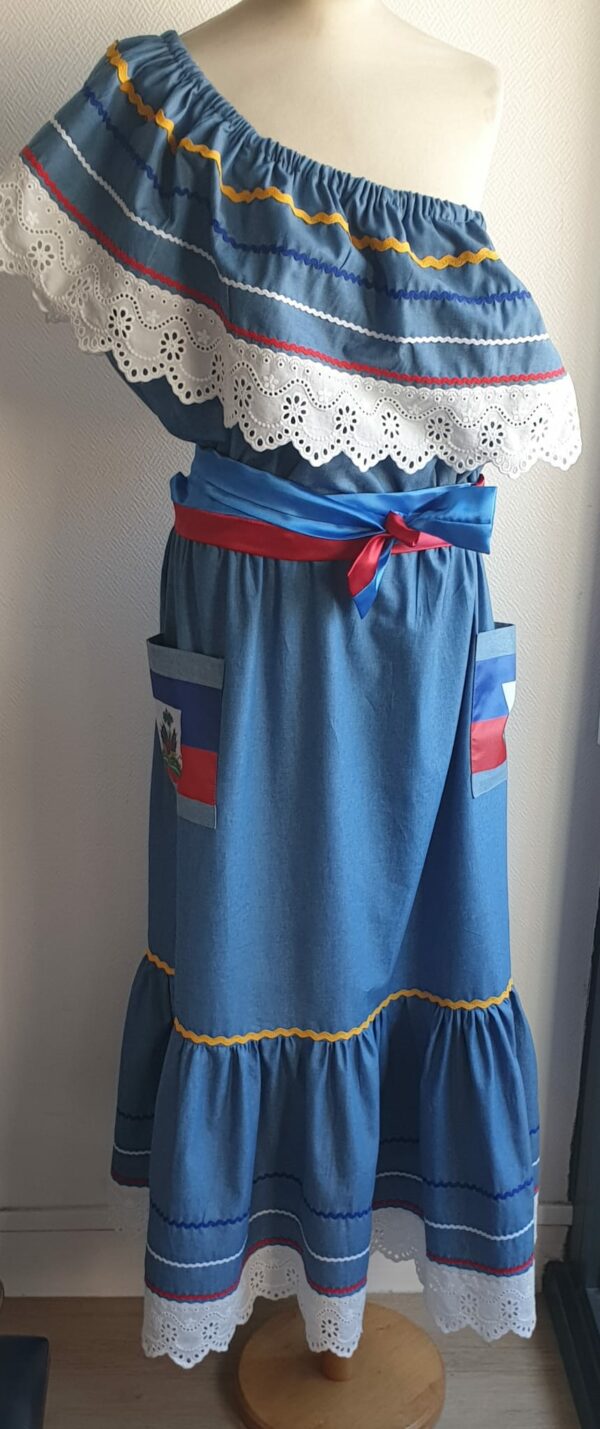 13.3Robe carabela longue en bleu sur une tenue traditionnelle. Le col bardot et le bas de robe sont brodés en dentelles animées. Les Zigzags sur le haut et le bas de robe sont respectivement jaunes, bleus foncé, blancs et rouges. Deux poches latérales rouges et bleues royales ornées du drapeau, ajoutent à l'harmonie du vêtement élastique à la partie rétrécie et 100% coton. Deux ceintures ont été pensées chacune d'entre elles en coureur unique, nouées à la taille. Entièrement confectionnée aux ateliers AnjéroCréation à Paris. Prix : 259.95€. 