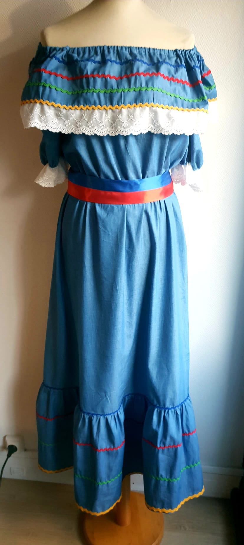 16.1Robe carabela en bleu clair sur une tenue traditionnelle aux manches courtes. Elle est composée entièrement de coton. Sa confection se distingue par une élasticité à la taille, un maxi col bardot et de broderies anglaises aux êxtrèmités.Les Zigzags en bas de robe sont proposés dans quatre couleurs. Cette robe est livrée avec une ceinture bicolore rouge et bleue. Toutes les tailles sont proposées. Prix : 199.95€. Entièrement conçu et réalisé dans les ateliers AnjéroCréation à Paris.