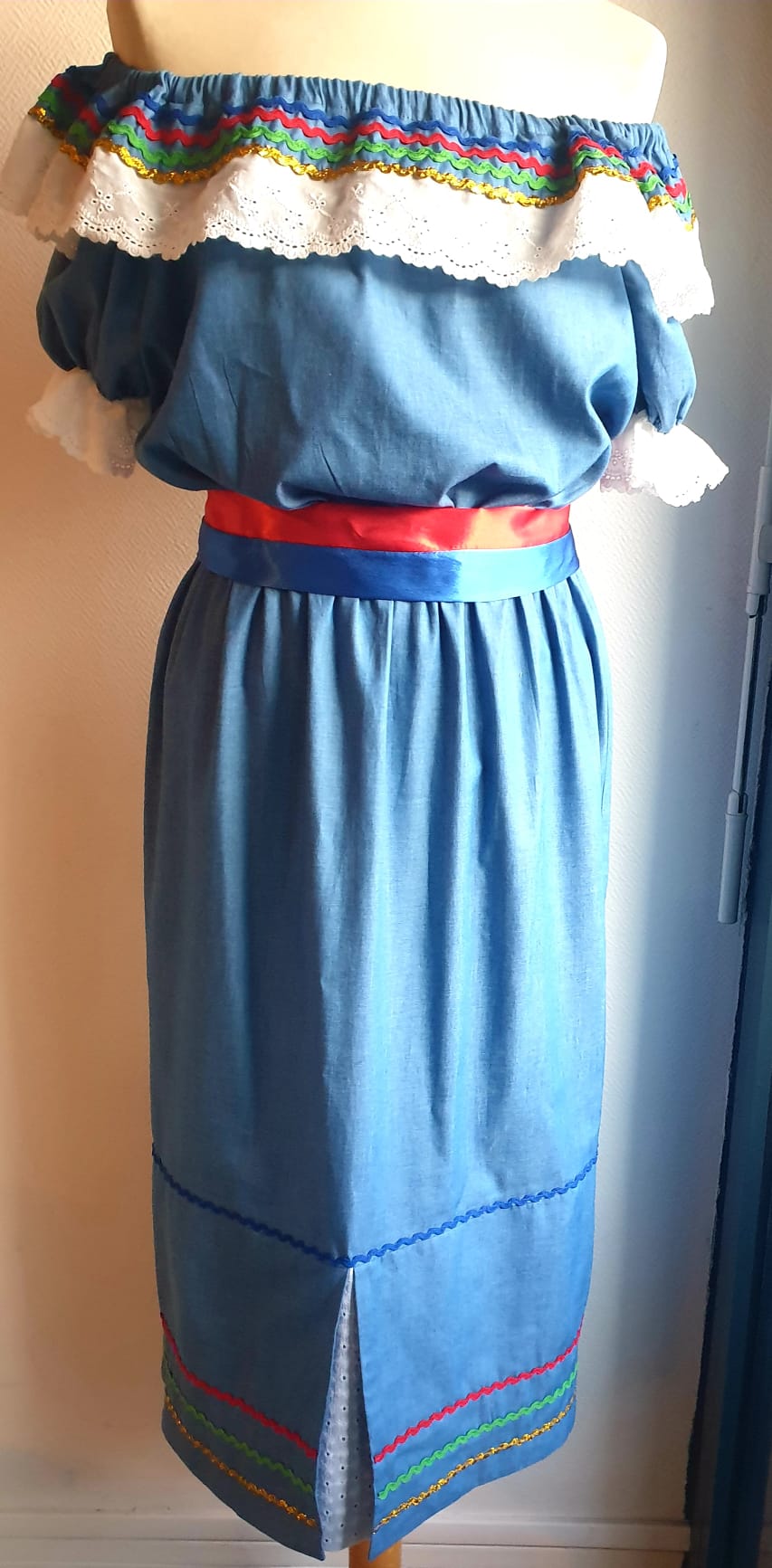 17.4Robe carabela en bleu clair sur une tenue traditionnelle aux manches courtes. Elle est composée entièrement de coton. Son style se distingue par une élasticité à la taille, un décoleté semi-plongeant, un maxi col bardot et de soyeuses broderies blanches anglaises aux êxtrèmités body. Les Zigzags ressérrés en bas et haut de robe proposés dans quatre couleurs de même que son fendu en son bas-milieu offrent ainsi une silhouette tout à fait originale. Cette robe est aussi livrée avec une ceinture bicolore rouge et bleue. Prix : 249.95€. Cette confection est entièrement conçue et réalisée dans les ateliers AnjéroCréation à Paris.
