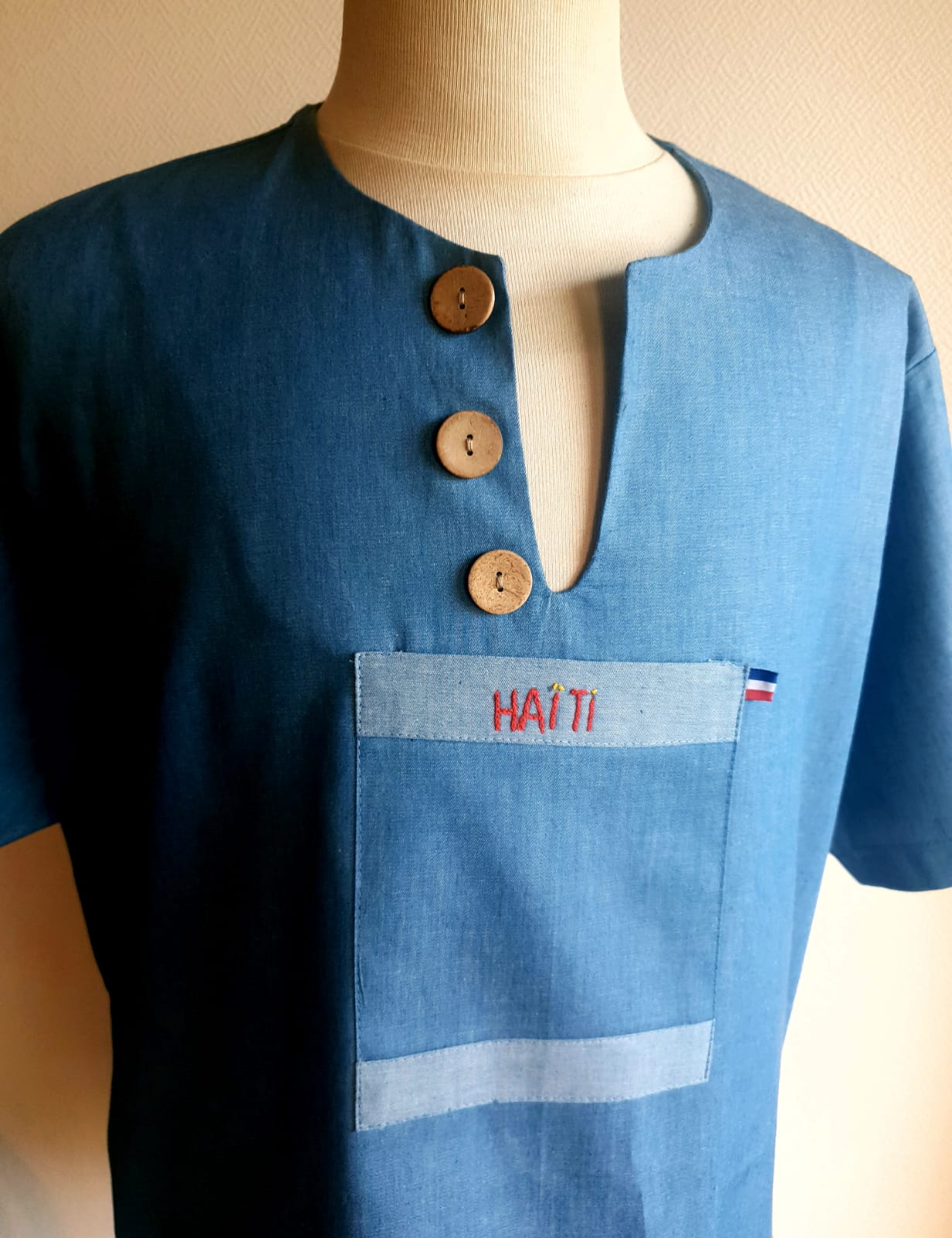1.1Chemise homme carabela en manches courtees et en coton sur une tenue traditionnelle haïtienne sur un ton bleu ciel. Un col ras du coup arrondi et fendu au devant duquel, trois petits boutons en bois coco sont proposés. Deux poches simples et une poche ornée d'une petite broderie Haïti confèrent un style original à cette chemise. Prix : 69.95€.