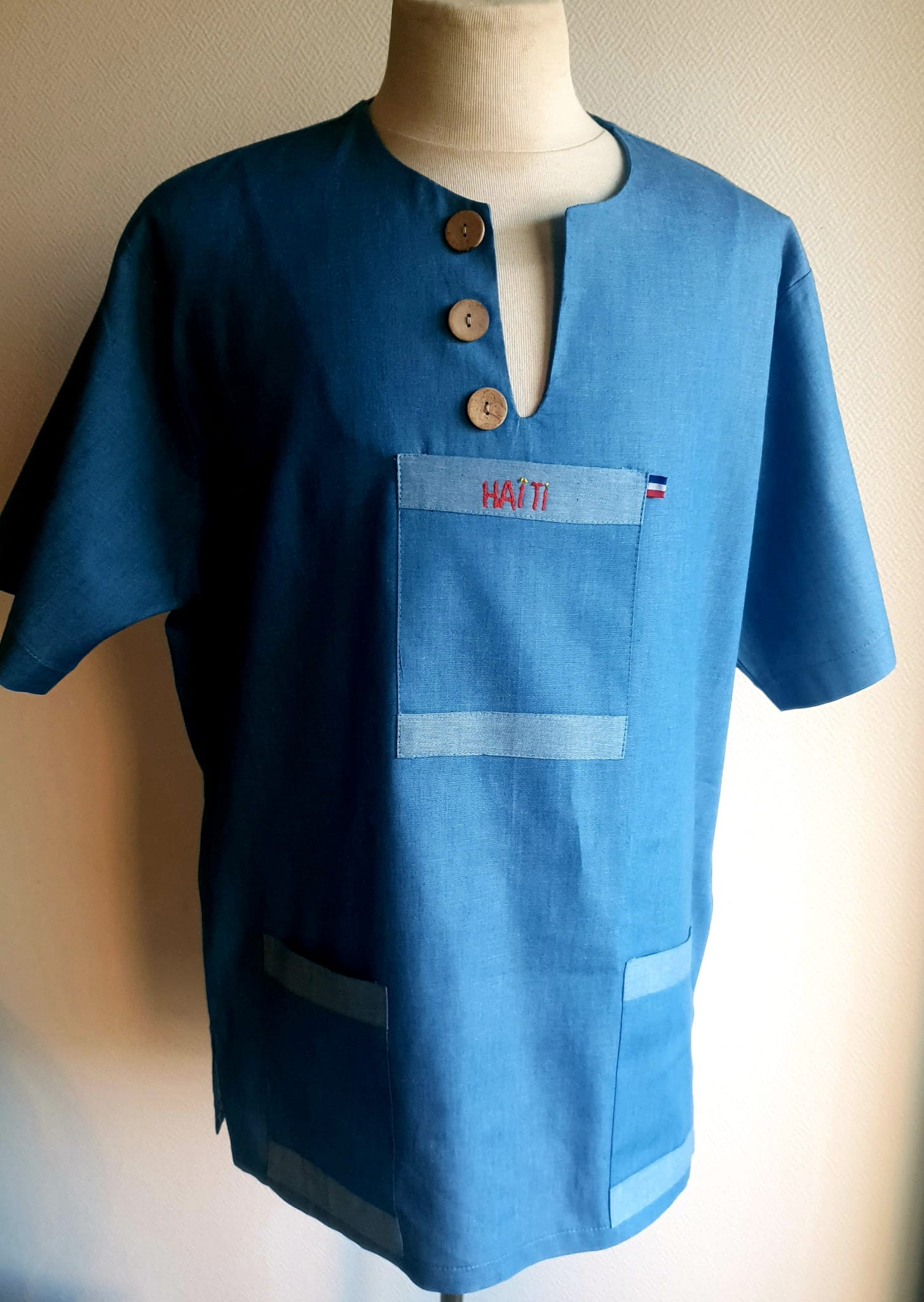 1.4Chemise homme carabela en manches courtees et en coton sur une tenue traditionnelle haïtienne sur un ton bleu ciel. Un col ras du coup arrondi et fendu au devant duquel, trois petits boutons en bois coco sont proposés. Deux poches simples et une poche ornée d'une petite broderie Haïti confèrent un style original à cette chemise. Prix : 69.95€.