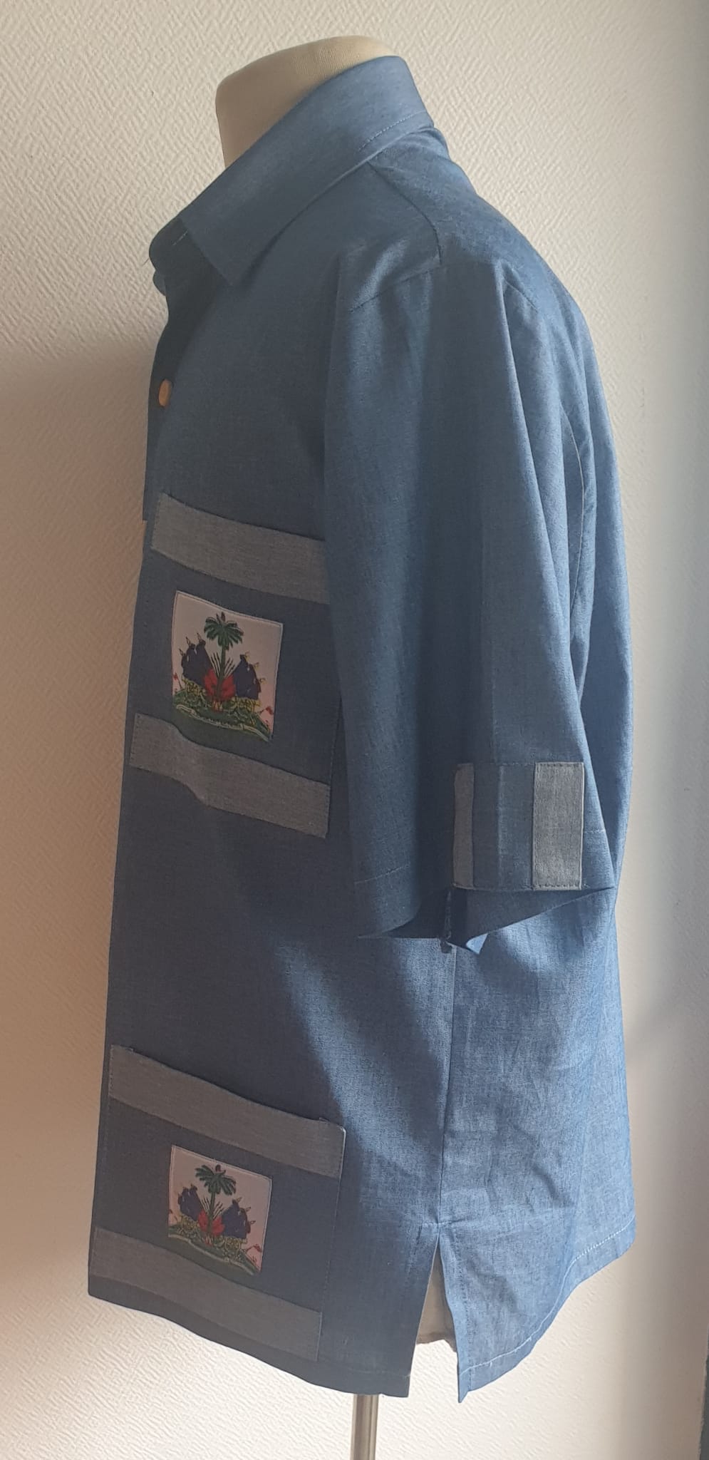 4.1Chemise carabela en tissu cent pour cent coton. Trois poches dont une munie du drapeau haïtien. Tissu travaillé à la main avec une petite poche cousue sur manche courte. Un Col classique a été choisi pour ce modèle avec un ensemble linéaire de boutons en bois coco verticaux. Cette chemise est entièrement conçue et réalisée dans les ateliers AnjeroCréation à Paris. Prix : 79.95€.