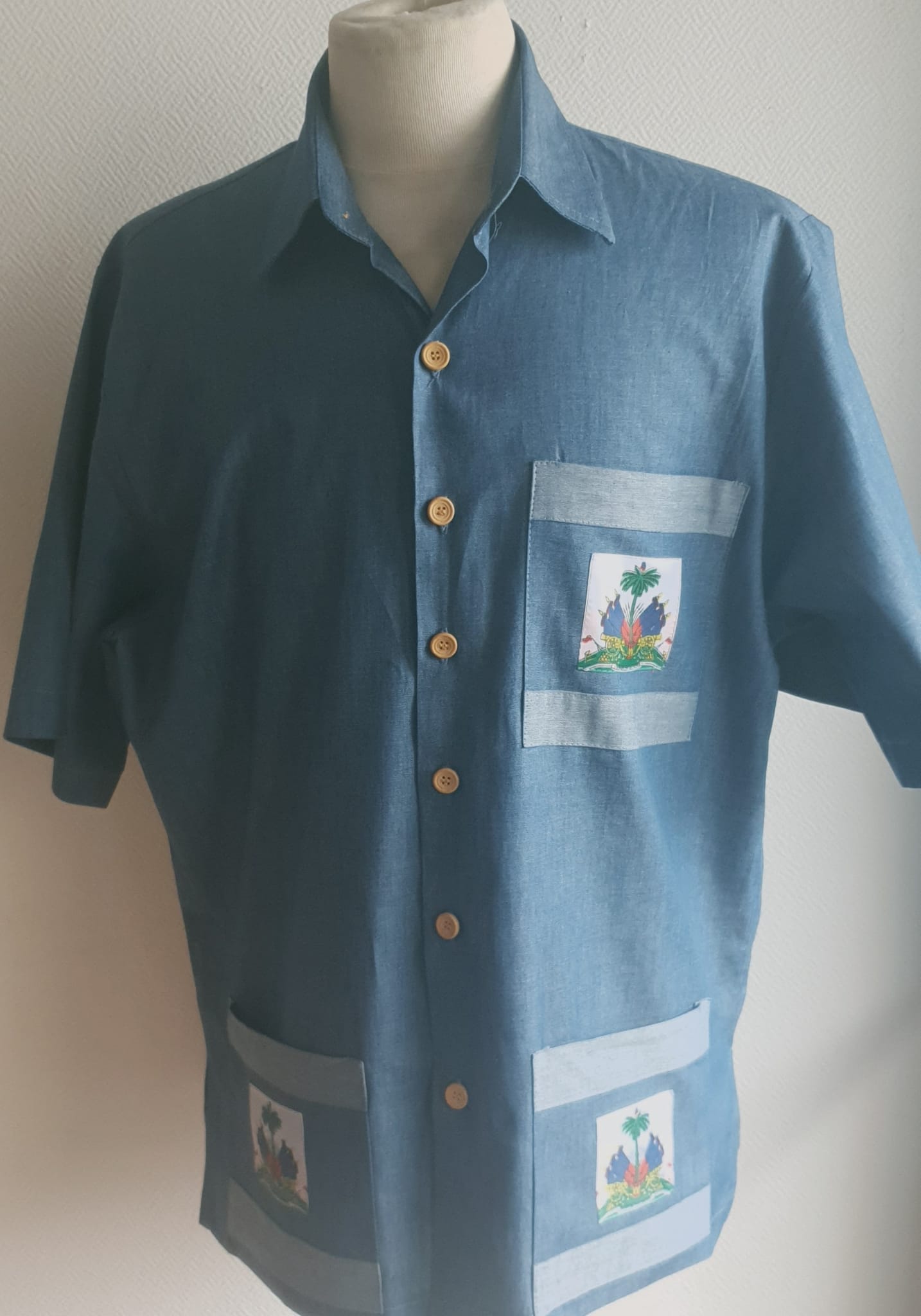 4.2Chemise carabela en tissu cent pour cent coton. Trois poches dont une munie du drapeau haïtien. Tissu travaillé à la main avec une petite poche cousue sur manche courte. Un Col classique a été choisi pour ce modèle avec un ensemble linéaire de boutons en bois coco verticaux. Cette chemise est entièrement conçue et réalisée dans les ateliers AnjeroCréation à Paris. Prix : 79.95€.