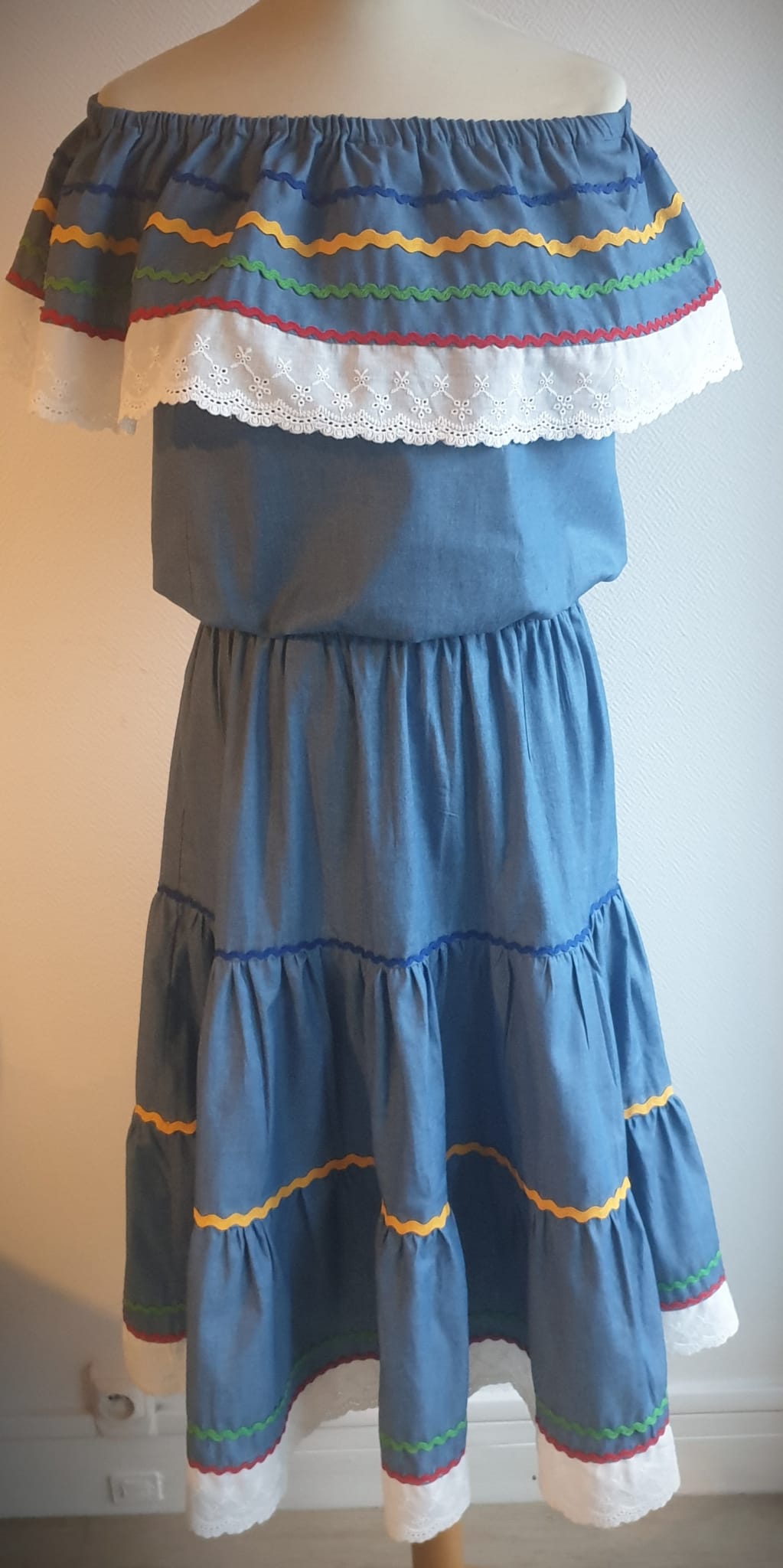 35.1Ensemble carabela bleu foncé sur une tenue traditionnelle haïtienne. Deux pièces. Jupe longue et élastique à la taille. Le corsage dispose au bas d'une appréciable élasticité, pouvant ainsi s'adapter à toutes les formes de buste. Les broderies blanches en dentelles anglaises sont cousues au bas du col bardot. Cette production est livrée avec, de surcroît, une grande ceinture aux couleurs du drapeau haïtien. Taille : S, M, L, XL, XXL. Prix : 169€. Création artisanale. Entièrement conçu et réalisé aux ateliers AnjeroCréation à Paris.