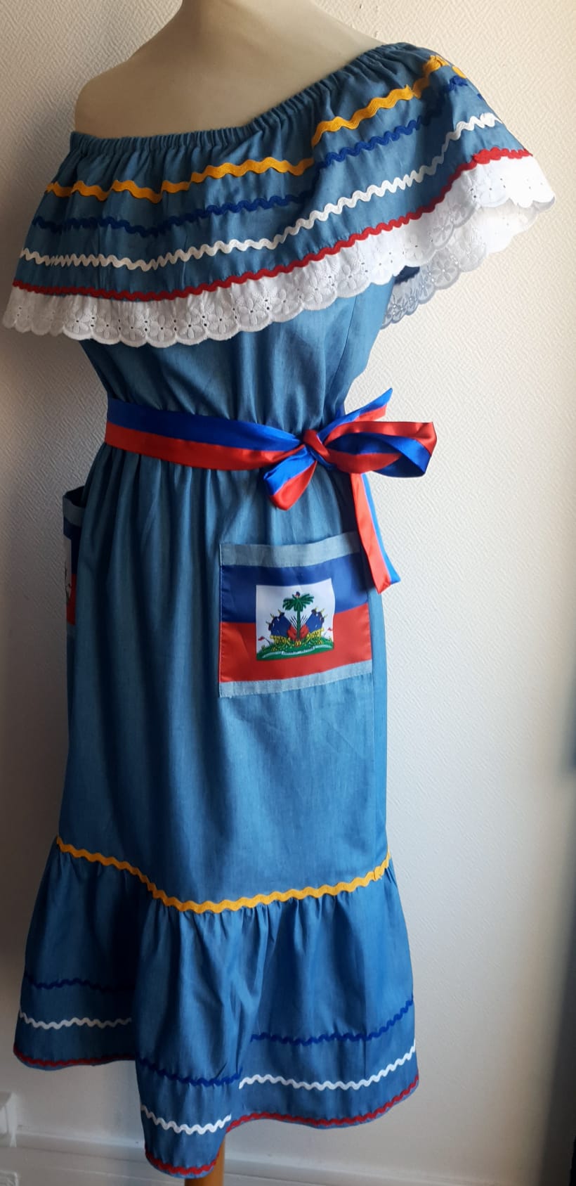 22.1 Robe Carabela mi-longue pour enfant sur une tenue traditionnelle haïtienne. Son bleu dominant et les serpentines décoratives découvrent un col bardot ajusté harmonieusement. Ce modèle cent pour cent coton laisse entrevoir de belles dentelles anglaises blanches en bas de robe. Taille : 12 ans. Prix : 149.95€. Ce produit est conçu et réalisé aux ateliers AnjeroCreation à Paris.