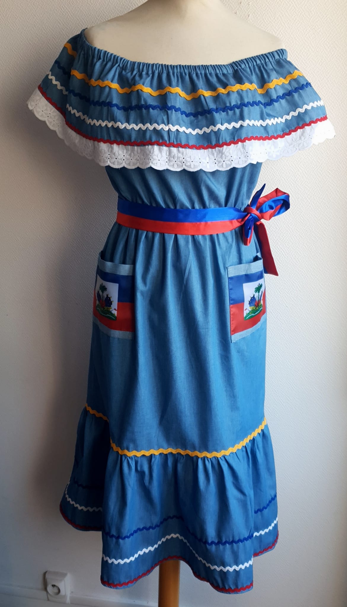 22.2 Robe Carabela mi-longue pour enfant sur une tenue traditionnelle haïtienne. Son bleu dominant et les serpentines décoratives découvrent un col bardot ajusté harmonieusement. Ce modèle cent pour cent coton laisse entrevoir de belles dentelles anglaises blanches en bas de robe. Taille : 12 ans. Prix : 149.95€. Ce produit est conçu et réalisé aux ateliers AnjeroCreation à Paris.
