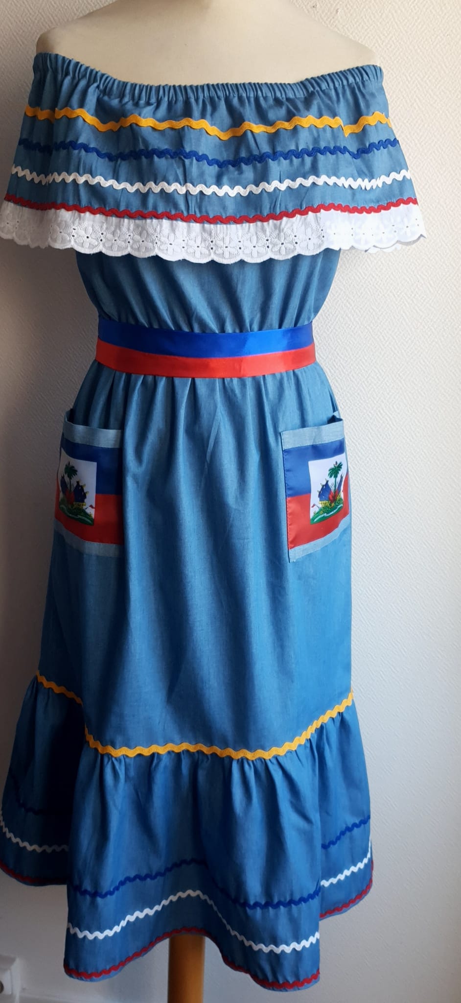 22.3 Robe Carabela mi-longue pour enfant sur une tenue traditionnelle haïtienne. Son bleu dominant et les serpentines décoratives découvrent un col bardot ajusté harmonieusement. Ce modèle cent pour cent coton laisse entrevoir de belles dentelles anglaises blanches en bas de robe. Taille : 12 ans. Prix : 149.95€. Ce produit est conçu et réalisé aux ateliers AnjeroCreation à Paris.