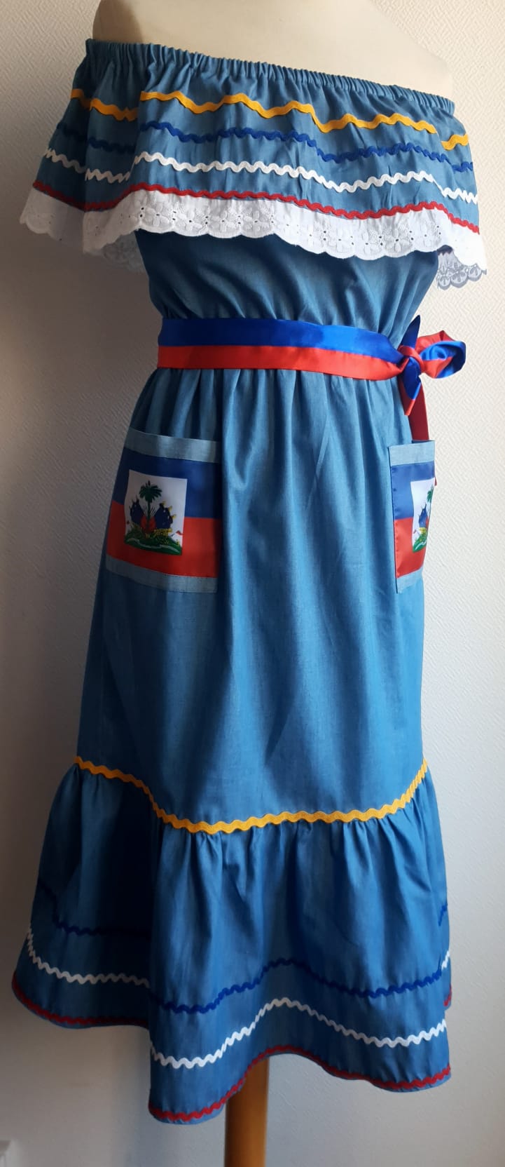 22.4 Robe Carabela mi-longue pour enfant sur une tenue traditionnelle haïtienne. Son bleu dominant et les serpentines décoratives découvrent un col bardot ajusté harmonieusement. Ce modèle cent pour cent coton laisse entrevoir de belles dentelles anglaises blanches en bas de robe. Taille : 12 ans. Prix : 149.95€. Ce produit est conçu et réalisé aux ateliers AnjeroCreation à Paris.