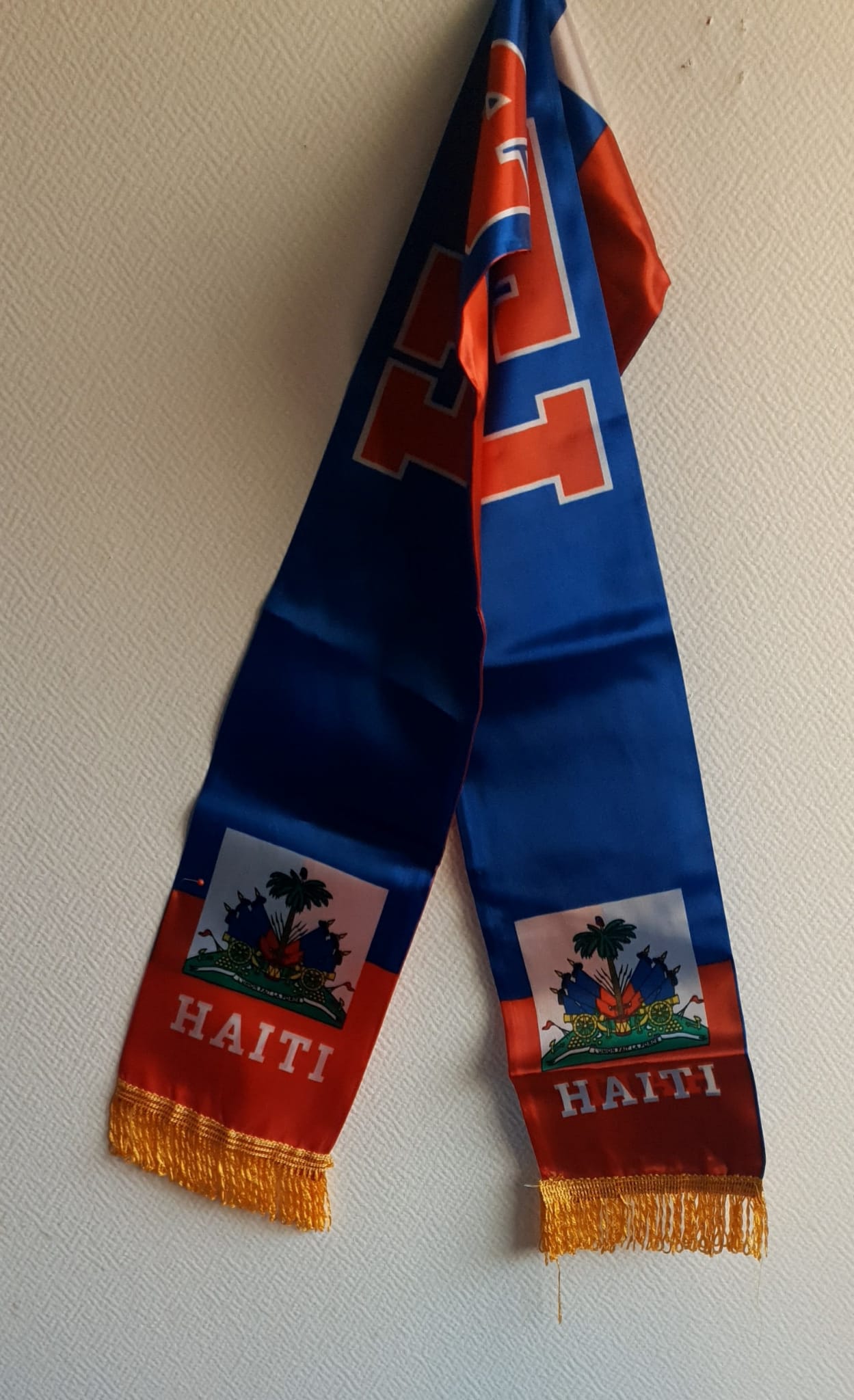 4.1Foulard réversible unisexe en satin bleuté. Des franges couleur or pensées aux extrémités pour la finition et la réversibilité des drapeaux haïtiens en décoration apportent une touche singulière et valorisent ce foulard. Dimension 1,35m en longueur sur 0,14m en largeur. Taille unique. Prix 7€.