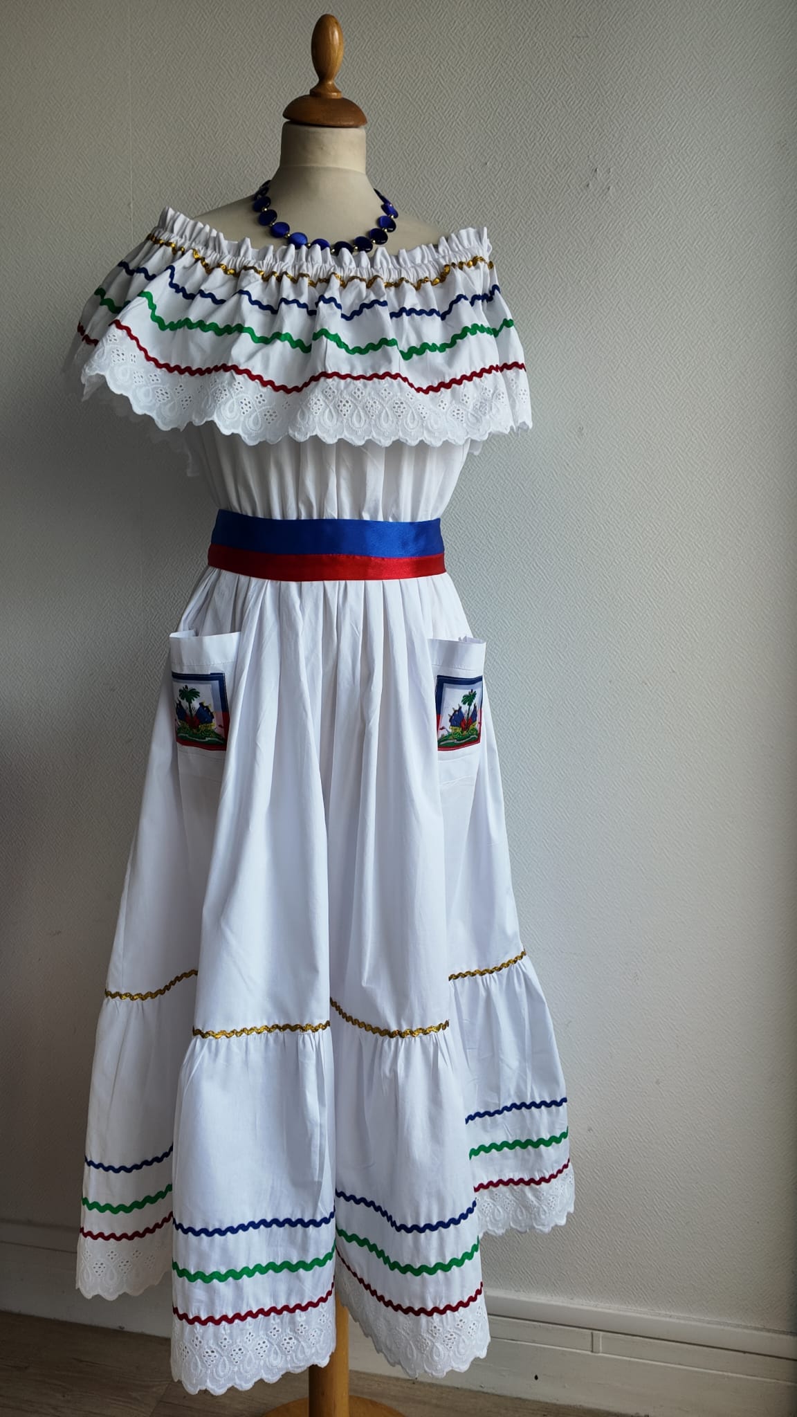 27.1Robe carabela en blanc, tissu popeline de coton. Élastique à la taille. Deux poches. Zigzags aux couleurs du drapeau haïtien. Dentelles décorées de broderies anglaises. La couleur de la robe fait merveilleusement bien ressortir la ceinture bleue, rouge. Peut être porté avec ou sans jupon. Modèle entièrement conçu et réalisé aux ateliers AnjeroCréation à Paris. Taille S, M, L, XL, XXL. Prix 359.95€