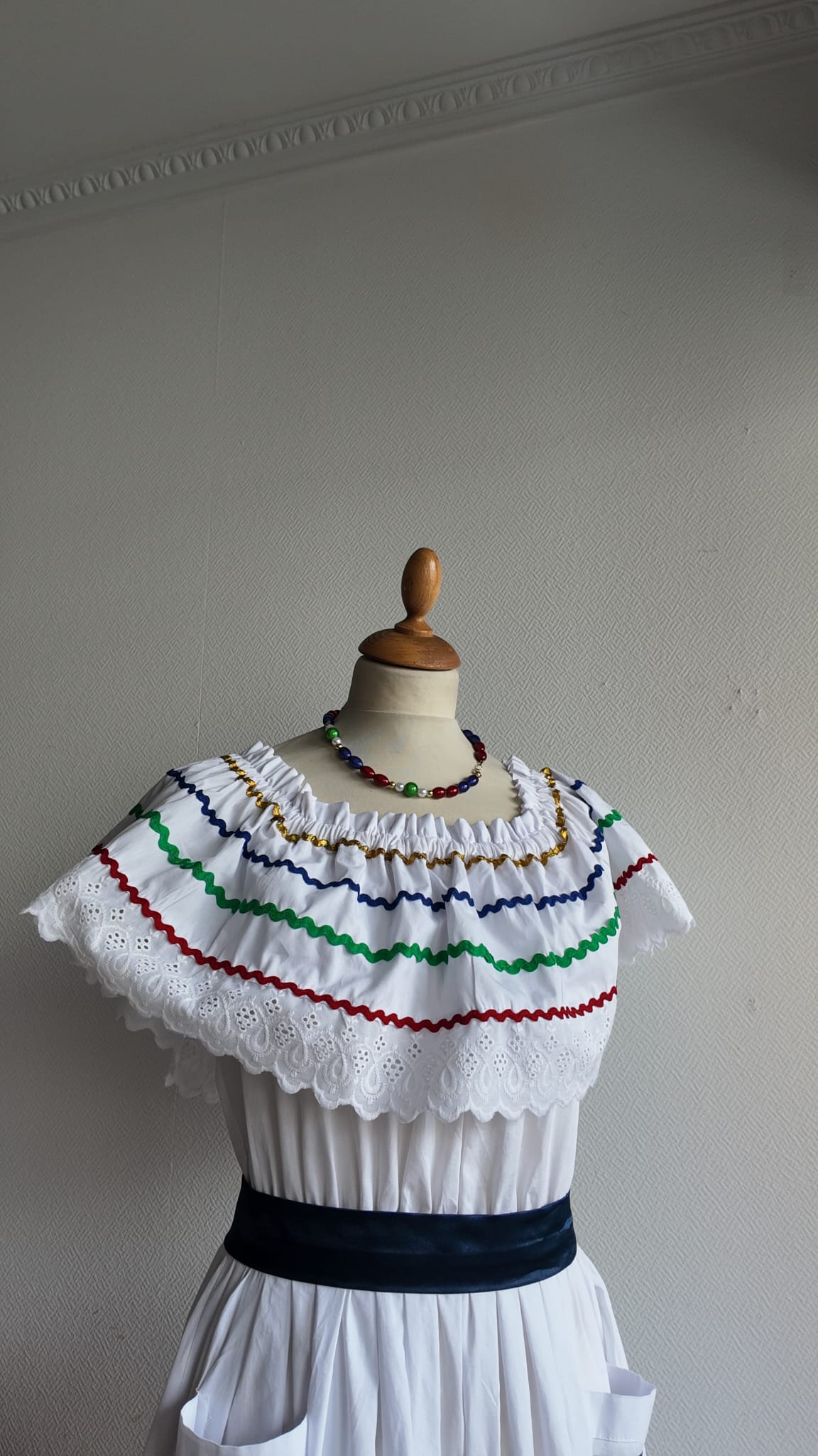 27.2Robe carabela en blanc, tissu popeline de coton. Élastique à la taille. Deux poches. Zigzags aux couleurs du drapeau haïtien. Dentelles décorées de broderies anglaises. La couleur de la robe fait merveilleusement bien ressortir la ceinture bleue, rouge. Peut être porté avec ou sans jupon. Modèle entièrement conçu et réalisé aux ateliers AnjeroCréation à Paris. Taille : S, M, L, XL, XXL. Prix : 359.95€