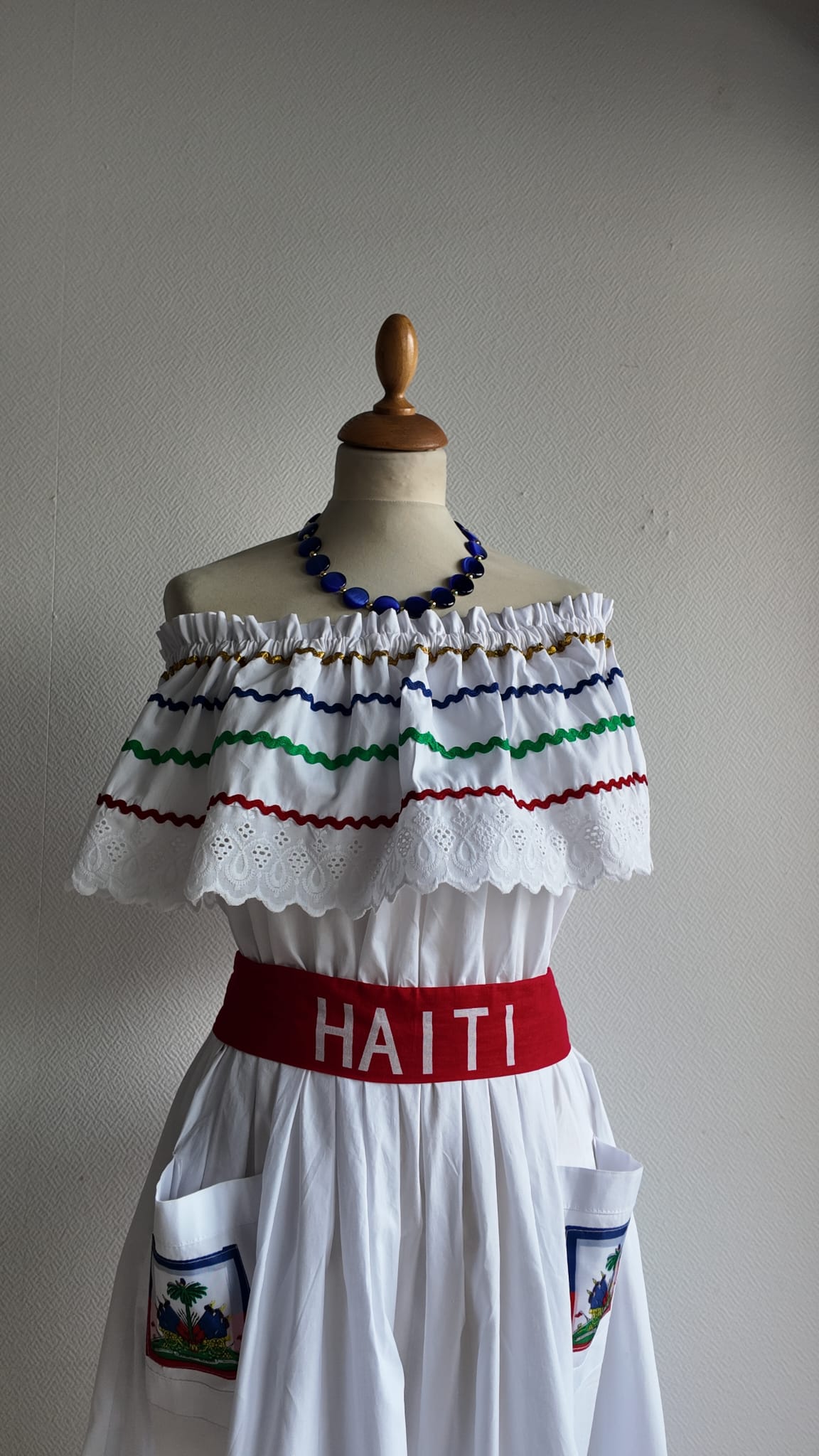 27.3Robe carabela en blanc, tissu popeline de coton. Élastique à la taille. Deux poches. Zigzags aux couleurs du drapeau haïtien. Dentelles décorées de broderies anglaises. La couleur de la robe fait merveilleusement bien ressortir la ceinture bleue, rouge. Peut être porté avec ou sans jupon. Modèle entièrement conçu et réalisé aux ateliers AnjeroCréation à Paris. Taille : S, M, L, XL, XXL. Prix : 359.95€