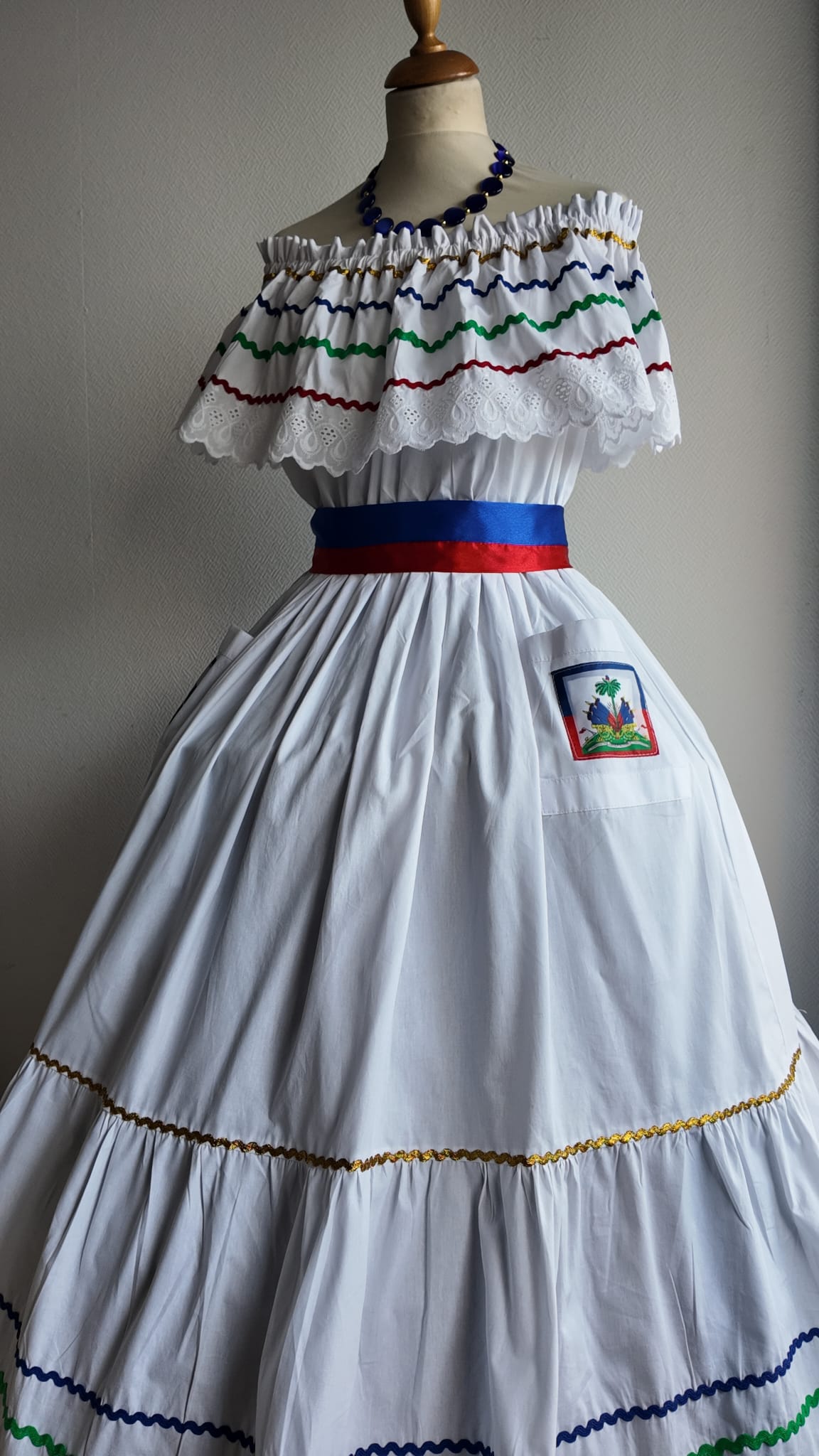 27.4Robe carabela en blanc, tissu popeline de coton. Élastique à la taille. Deux poches. Zigzags aux couleurs du drapeau haïtien. Dentelles décorées de broderies anglaises. La couleur de la robe fait merveilleusement bien ressortir la ceinture bleue, rouge. Peut être porté avec ou sans jupon. Modèle entièrement conçu et réalisé aux ateliers AnjeroCréation à Paris. Taille : S, M, L, XL, XXL. Prix : 359.95€